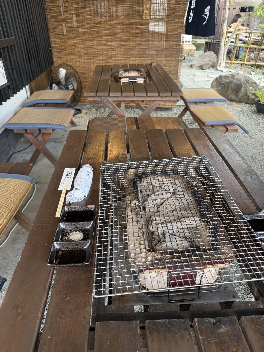 A heavy wooden table with a charcoal pit in the centre covered by a wire grill, dipping sauces in small black dishes beside it, semi-outdoor seating on gravel behind